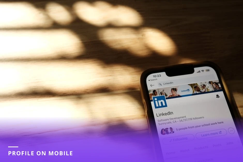 Career coach at a bright modern Western office desk looking down at a smartphone showing a LinkedIn profile, laptop open and notebook beside them, natural window light, candid focus