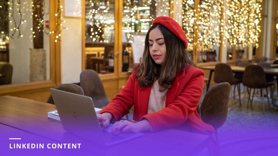 Woman in a bright Scandinavian-style cafe typing on a laptop with a notebook, pen, coffee cup, and smartphone showing a social feed beside her.