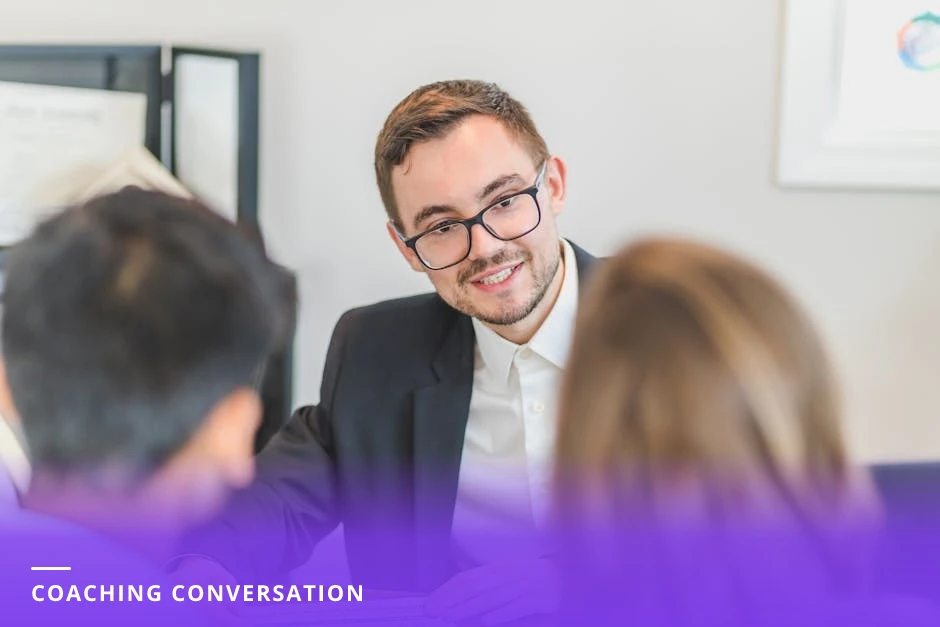 Two professionals in a European-style office cafe: a career coach leaning forward, gesturing while the client listens attentively across a small table, warm natural light