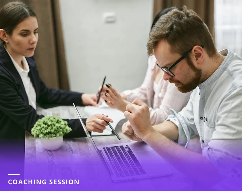 Coach and client leaning over a notebook and laptop at a table in a modern European office, engaged in a focused one-on-one discussion.