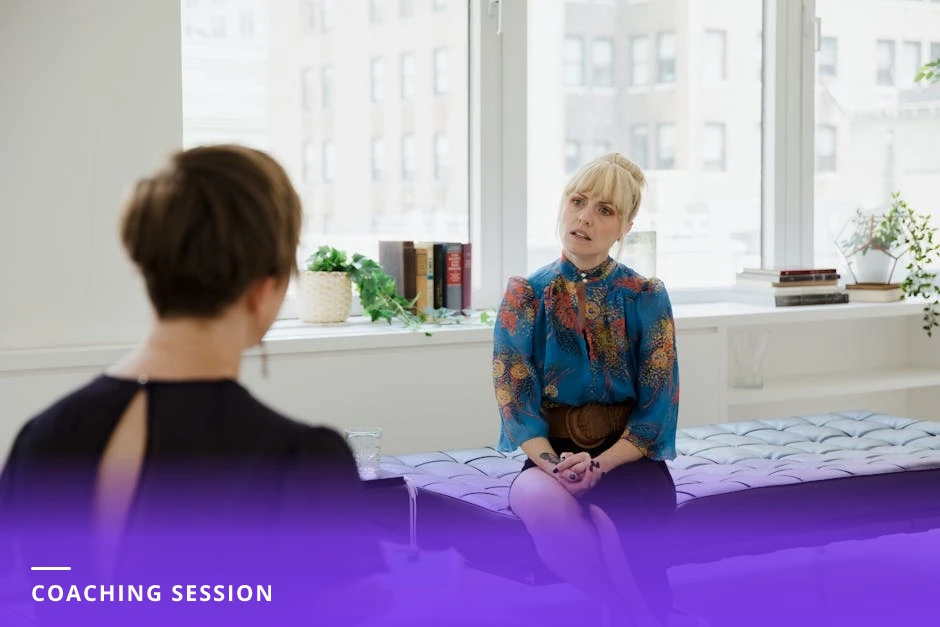 Candid one-on-one coaching conversation in a bright modern Western office, coach listening and taking notes while client speaks across a small table.