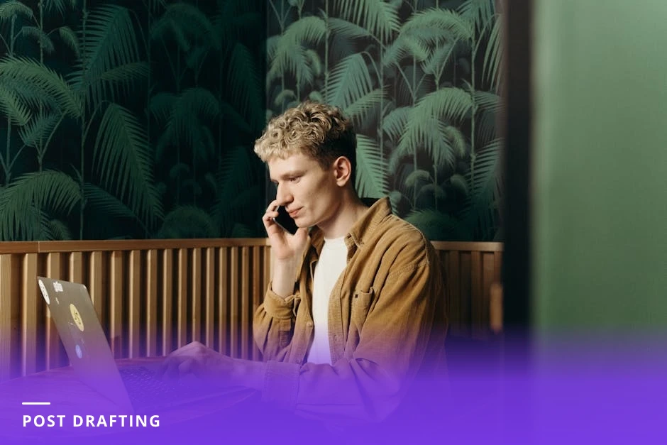 Close-up of a young professional's hands typing on a laptop at a cafe table with a smartphone displaying a LinkedIn post draft, notebook and coffee nearby.