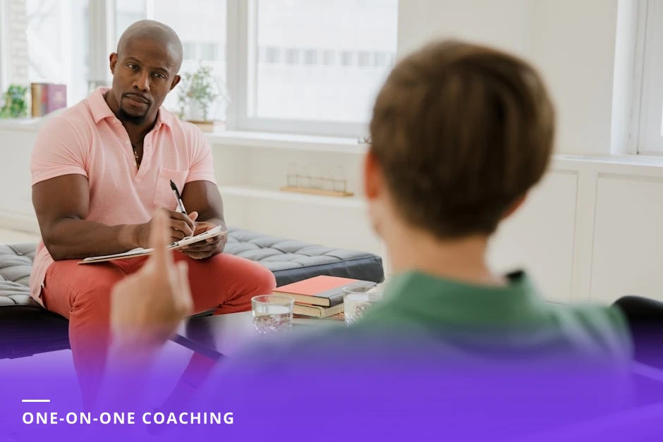 Coach and client in a bright European office, coach listening and writing in a notebook while the client gestures, candid supportive conversation