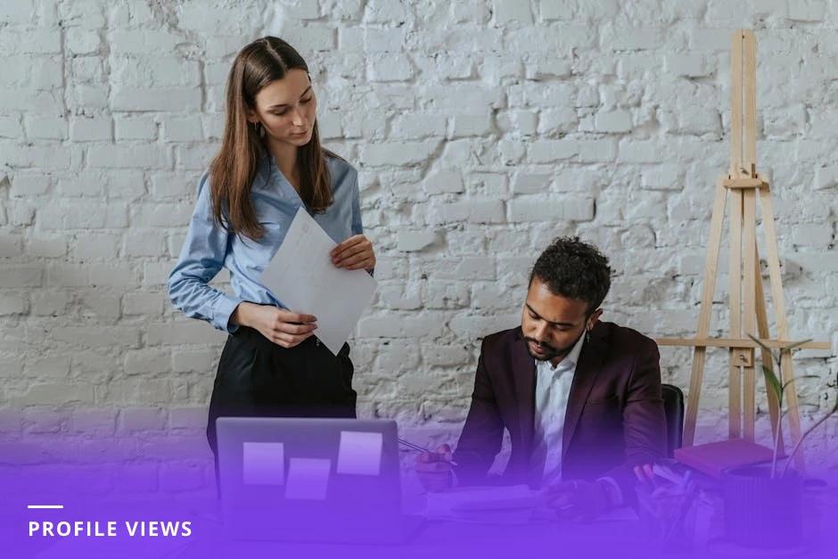 Professional coach at a bright Scandinavian-style office desk, viewing a laptop screen that shows LinkedIn-style analytics charts (profile views, follower demographics), taking notes in a notebook.