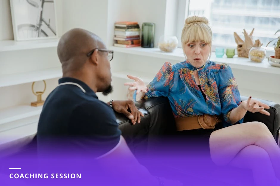 A coach and client in a bright modern Western office having a candid one-on-one conversation across a small table; coach leans forward, attentive, taking notes while client gestures, natural light through windows.