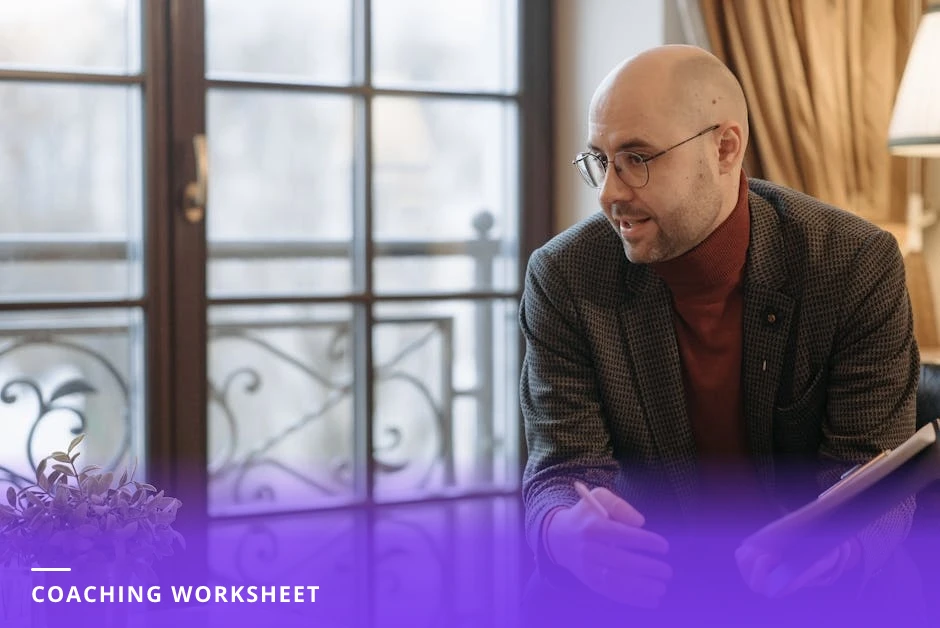 A diverse coach and client sit across a wooden table in a bright Scandinavian-style office reviewing a single-page worksheet; coach points to questions while a laptop, notebook, and coffee cup sit nearby.