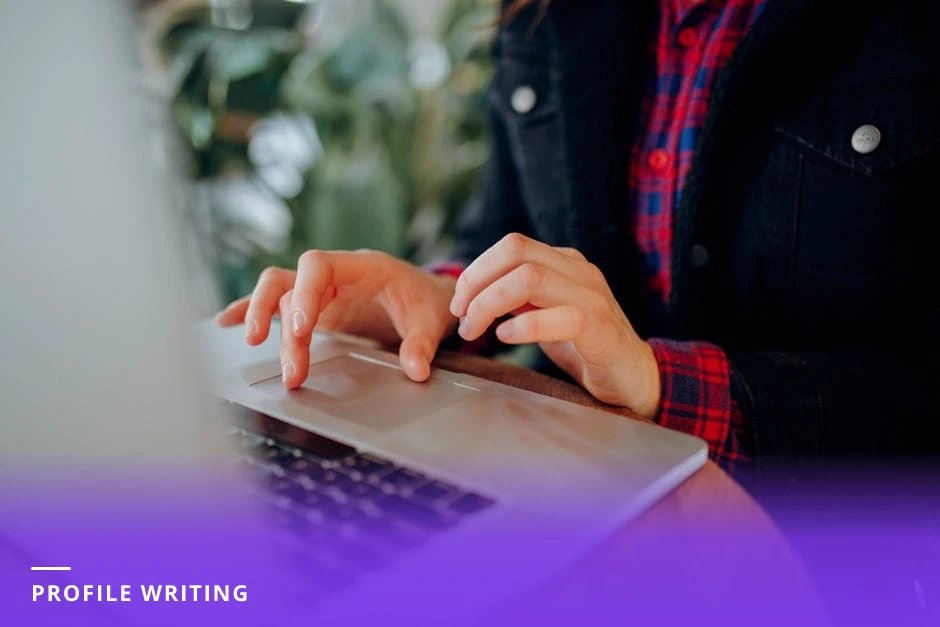 Mid-career professional typing on a laptop at a sunlit cafe table, coffee beside their notebook, focused expression as they edit a profile or About section