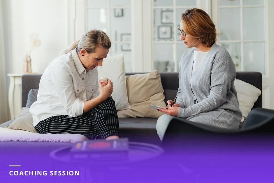 Coach sitting across from a client in a bright Scandinavian-style office, leaning forward with a notepad while the client speaks, natural light and plants in background