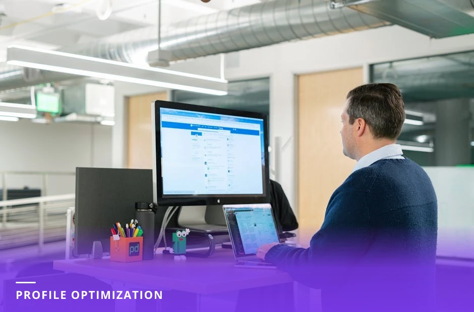 Coach at a modern Western office desk in natural light, focused on a laptop displaying a LinkedIn profile with notebook and coffee nearby.