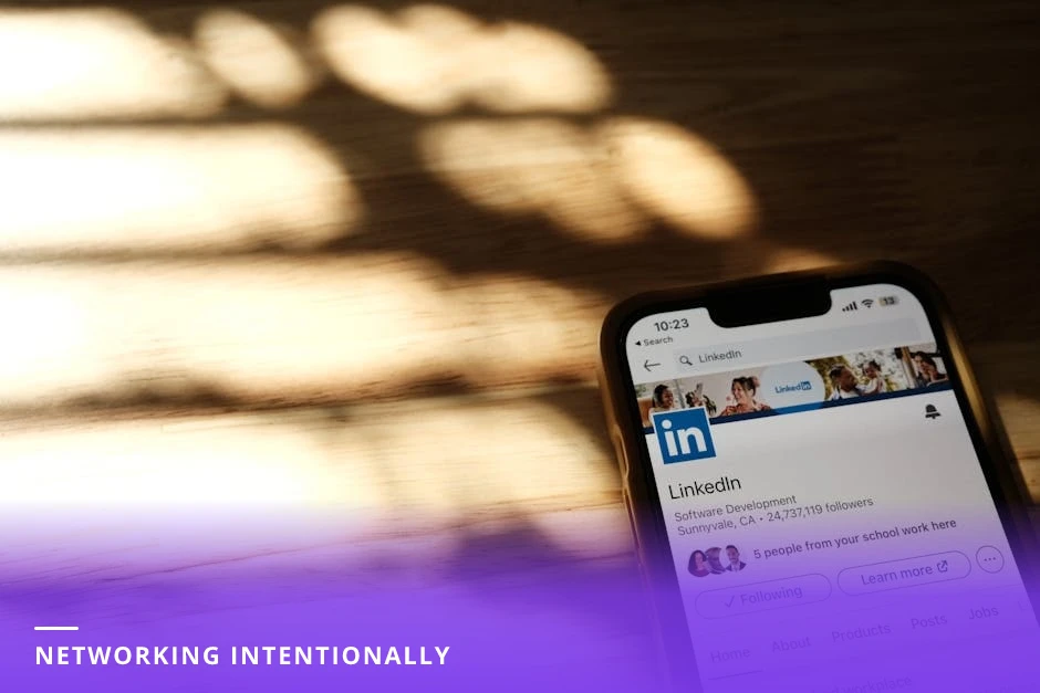 Two professionals at a small table in a European cafe, leaning in and reviewing a phone showing a LinkedIn profile, candid conversation in natural light.