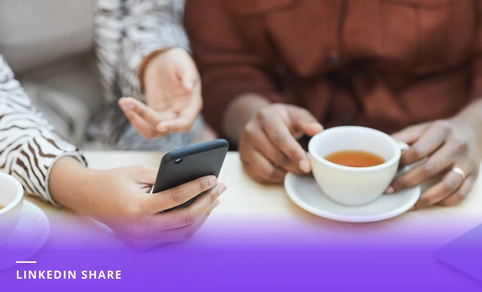 Two colleagues at a cozy cafe table, one holding a smartphone showing a LinkedIn profile while the other leans in, coffee cups and notebook on table