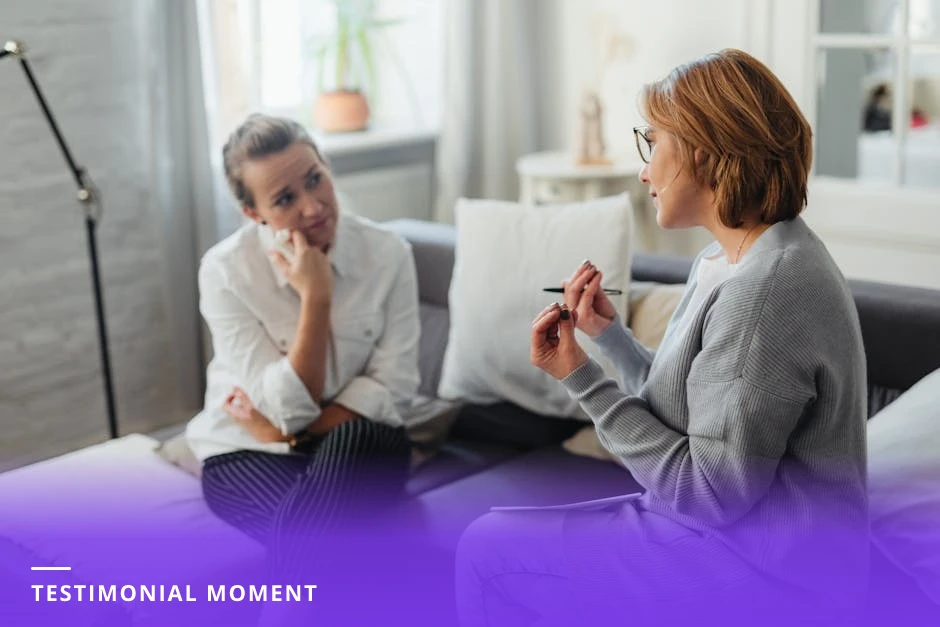 A coach and client sit across a small table in a modern Western office; the client is tearful and speaking while the coach leans forward listening attentively, notebooks and a glass of water on the table, soft natural light