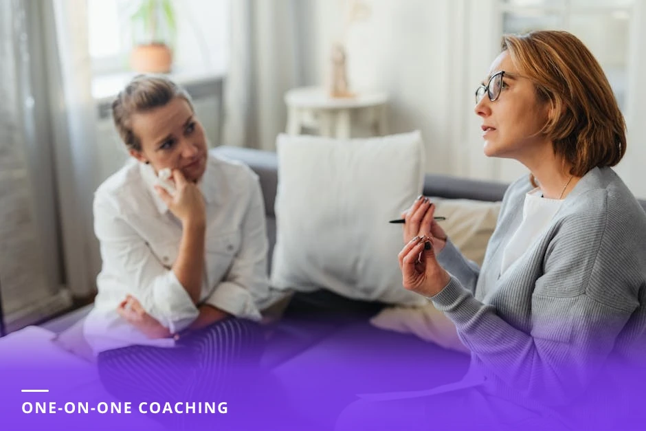 A career coach leans forward listening attentively to a client across a wooden table in a sunlit modern cafe; laptop, notebook, and coffee cups on the table.