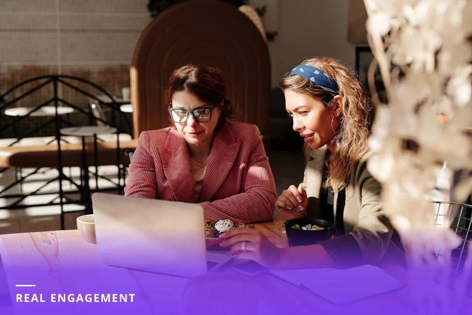 Two professionals conversing across a small table in a bright modern Western coffee shop, leaning in and smiling during a candid networking exchange.