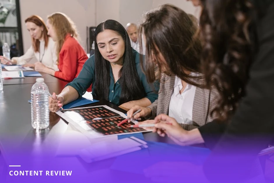 Two diverse professionals at a cafe table leaning over printed slide mockups and sticky notes, one pointing at a slide while the other takes notes, phones and pens on the table