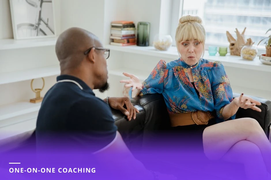 Coach and client sit across a small table in a bright modern European office; the coach leans forward listening and taking notes in a notebook, natural light through large windows, laptop and coffee on the table