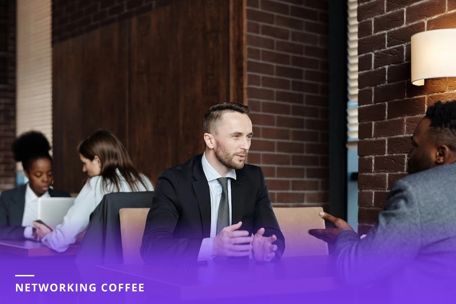 Two diverse professionals in business casual talking across a cafe table with laptops and coffee, candid networking conversation in natural light.
