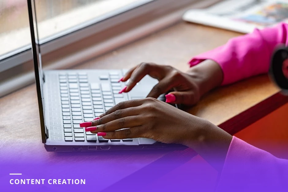 Young professional woman typing on a laptop with a smartphone nearby showing a LinkedIn profile; bright Scandinavian-style workspace with coffee mug and sticky notes on desk.